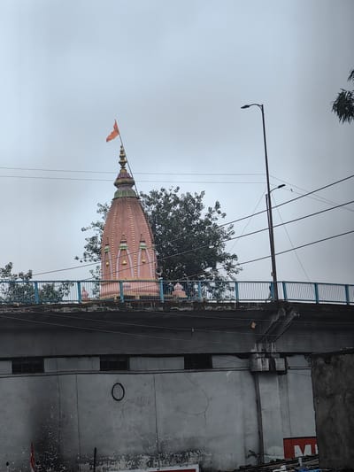 A pink temple spire, topped with a golden finial and an orange flag, rises against a hazy sky. The spire is decorated with light green accents and small bell-shaped ornaments. It appears to be situated on a raised platform or overpass, indicated by a blue metal railing and a concrete structure beneath it. A tall street lamp with a dark fixture stands to the right, its pole partially obscured by power lines crisscrossing the frame. Trees with dark foliage are visible behind the temple spire and to the left. The lower portion of the image shows a grey concrete wall with several rectangular openings and a circular marking. In the foreground, some debris and possibly parts of signs are visible. This scene is located near the historic Gohar Mahal in Bhopal, India, which is situated on the banks of the Upper Lake.