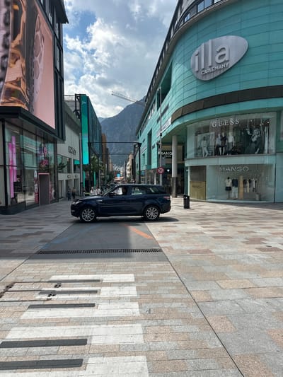 The image shows a street in Les Escaldes, Andorra. A dark blue Range Rover is parked on the right side of the street, casting a shadow. Buildings flank both sides of the street. On the left is a building with a digital advertising display and various shops. The right side features storefronts for Guess and Mango. Above the Guess and Mango shops, "ILLA CARLEMANY" is visible on a building. A mountain range is visible in the distance. The street is paved with gray and beige stones arranged in a linear pattern. Pedestrians can be seen walking on the sidewalk. A street sign is partially visible.