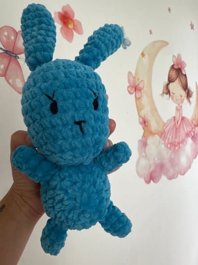 The image shows a close-up of a handmade blue crocheted bunny being held by a person's hand. The bunny has black eyes and a small black stitched mouth and nose. In the background, there is a white wall decorated with decals. The decals include a pink butterfly, a pink flower, a crescent moon with a star, and a drawing of a young girl with pigtails in a pink tutu sitting on clouds. The scene appears to be indoors, possibly a child's bedroom, and there is no indication of the location being in the city of Oldham, United Kingdom.