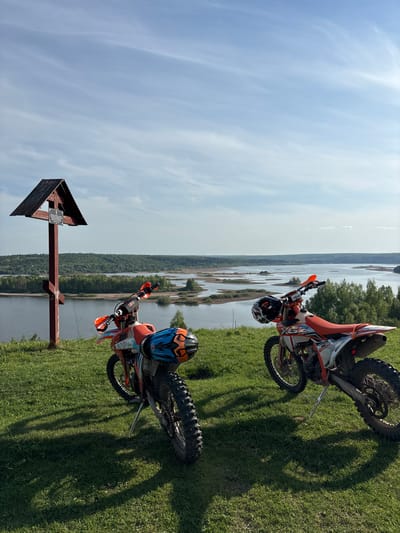 Two orange and white dirt bikes are parked on a grassy hill overlooking a body of water. A weathered wooden arrow-shaped signpost with a small plaque is present in the foreground. The background shows a landscape of water, trees, and sky, consistent with an open natural area. The scene is Serga, Russia.
