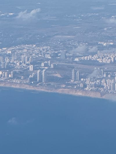 This is an aerial photograph of a coastal city with a beach and the sea. The image is oriented vertically, with the city and beach on the left and the sea on the right. The city appears densely populated with numerous buildings, including what look like apartment complexes and other structures. The buildings are clustered together, and some are of a modern architectural style with multiple stories. The beach is a strip of sand bordering the city, and beyond that is the vast expanse of the ocean. The water is a deep blue, suggesting it is a clear day. Some cloud formations are visible in the sky, as well as in the distance over the city. The lighting suggests it is daytime, likely with good visibility. The overall scene depicts an urban environment situated along a natural coastline. The image has a slight haze, which may be due to atmospheric conditions or the altitude from which the photograph was taken. There is no visible text in the image.