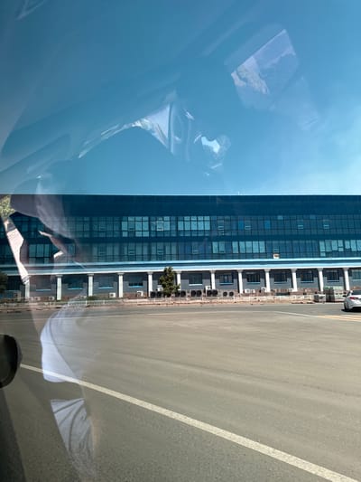 The image shows a modern building with a dark blue glass facade, reflecting the sky and surrounding environment. The building has multiple stories with a grid of windows. Below the glass facade, there is a lighter-colored section with smaller windows and air conditioning units installed on the exterior. A paved area, likely a parking lot or road, is in the foreground, with white lines marking lanes. A white car is visible parked on the right side of the frame. The reflection on the left side of the image suggests the photo was taken from inside a vehicle, possibly with the windshield or side window reflecting the interior and the exterior scene. The sky is a clear blue. This scene is likely located in Jezqazğan, Kazakhstan, given the prompt's instruction, but this cannot be visually confirmed from the image alone.