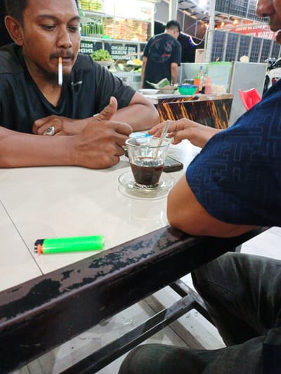 This media file shows two individuals in what appears to be an outdoor eatery or cafe, likely in Cotpuuk, Indonesia. The lighting suggests it could be daytime or early evening.
In the foreground, a man with a mustache is seated at a table, smoking a cigarette. He is wearing a black t-shirt and a ring on his left hand. His arms are resting on the table, and he is looking down. Next to him, a glass of dark liquid, presumably coffee, sits on a saucer with a spoon. Also on the table is a bright green lighter.
The background reveals more of the setting. Another person, wearing a black t-shirt with a graphic design, is standing and looking away from the camera. Shelving units stocked with various items, possibly food or supplies, are visible behind him. There are also some plastic containers and bottles on a counter. The overall impression is a casual, open-air establishment.
The image captures a moment of leisure or a brief stop at a local spot. The focus is on the two men and the immediate surroundings of their table.