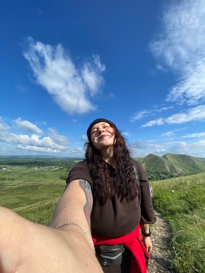 A woman with long, dark, curly hair, wearing a black beanie, a brown sweatshirt, and a red garment tied around her waist, is captured in a selfie. Her eyes are closed, and her face is tilted upwards, seemingly enjoying the sun. She has a tattoo on her left arm and is wearing a silver bracelet. The woman is standing on a gravel path on a grassy hillside. In the background, a rural landscape stretches out, featuring green fields, a village with scattered houses, and rolling, tree-covered hills under a bright blue sky with scattered white clouds. The location is the city of Andreevka, Russia.