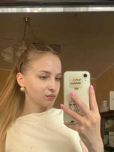 The image is a vertical, self-taken photograph of a young woman in Kyiv, Ukraine. She is holding a smartphone with a white case up to a mirror, capturing her reflection. The woman has light-colored hair and fair skin with freckles. She is wearing a white top and has a pair of wire-rimmed sunglasses resting on her head, pushed back from her face. She is also wearing a large, textured gold earring in her visible ear. Her fingernails are painted a bright pink.
On the back of her phone case, a square sticker with black handwritten text is visible. The text is written in Cyrillic script and reads "НЕ ВОЖЖЯ" which translates to "Not a whip." Below the text is a simple, cartoonish drawing of a face with large eyes and a small mouth.
The background of the image is blurry but appears to be an indoor setting. The wall behind her is a neutral, light brown color. A reflection in the mirror shows what appears to be a window with vertical blinds and possibly shelves with boxes in the upper background, suggesting a home or office environment. The lighting in the photo is soft, indicating it was likely taken during the daytime. The woman's expression is neutral, and the overall mood of the image is casual and everyday. There are no explicit indicators of the weather.