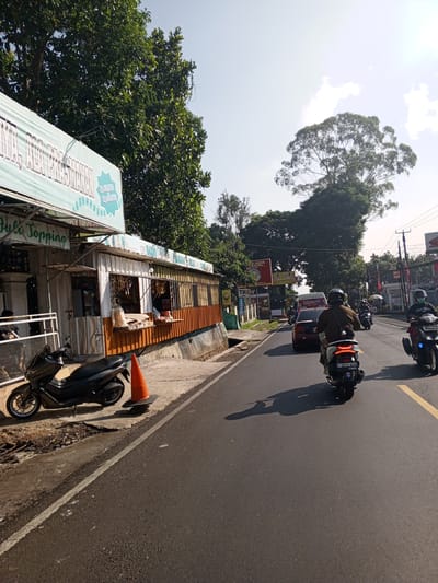 This image depicts a sunlit street scene in Weddagala, Sri Lanka, characterized by a mix of commercial establishments, dense vegetation, and ongoing road traffic. The time of day appears to be late morning or early afternoon, indicated by the clear blue sky with sparse white clouds and the distinct, elongated shadows cast by vehicles on the asphalt. On the left side of the frame, a single-story commercial building, possibly constructed from corrugated metal or similar materials, features an open storefront. An awning above the shop entrance displays a light blue banner with white text, partially legible as "Hulo Topping" and "LA PRAWANAN". Another section of the banner shows pricing in Indonesian Rupiah: "Rp.15.000" and "Rp.20.000". Goods, likely packaged snacks, are displayed on shelves within the open shop window. A person is visible inside the shop, possibly attending to customers. Parked on the concrete sidewalk in front of this shop is a black Yamaha NMAX scooter, facing left. An orange traffic cone is positioned on the sidewalk near the scooter. Further down the street on the left, additional signs are visible, including one with the text "ALAM SUNDA", and dense green foliage lines the roadside. The asphalt road, which appears dry, occupies the mid-ground and extends into the distance. It features a solid white line marking the edge and a broken yellow line delineating lanes. Traffic flows on the right side of the road. In the immediate foreground on the road, a person riding a white scooter is seen from behind, wearing a brown jacket and a dark helmet. The scooter's license plate, "F 2878 WAH," is clearly visible. To the right of this scooter, another person on a black scooter is also seen from behind, wearing a dark jacket, a dark helmet, and a face mask. Its license plate is partially visible as "D 6165 MAJ." Further up the road, a dark red sedan is visible behind the white scooter, and a white van or bus is partially seen behind the car. Several other motorcycles and distant vehicles are also present. The right side of the image is dominated by lush, green trees and vegetation. A particularly large, mature tree with a broad canopy stands prominently against the bright sky in the upper right quadrant. Utility poles with overhead wires run along the right side of the road. In the distance, various commercial signs, including one for "Honda," are discernible