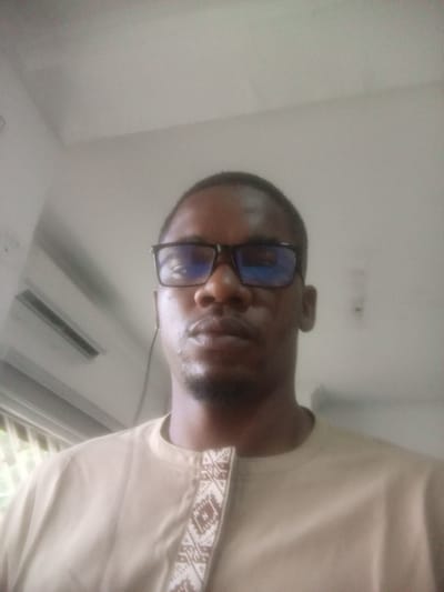 A medium shot photograph shows a person with dark skin, wearing glasses and a light-colored garment with a patterned neckline. The person is looking directly at the camera. The background is a plain, lightly colored wall. The lighting is soft and diffused, suggesting an indoor setting. There are no discernible city landmarks or any indication of location in Sudbury, United States of America.