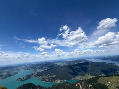 This is a high-angle aerial photograph of a vast landscape with a bright blue sky filled with scattered white clouds. The scene depicts rolling green hills and mountains covered in dark green forests. A large, turquoise lake dominates the foreground and middle ground, with several peninsulas and inlets. Along the shores of the lake, small villages and scattered houses are visible. In the distance, more mountain ranges and bodies of water stretch towards the horizon. The image appears to have been taken from a high vantage point, likely a mountain peak, overlooking the Wolfgangsee lake and surrounding area near Sankt Gilgen, Austria. There are no people or animals visible in the photograph. The overall impression is one of a serene and expansive natural landscape on a clear day.