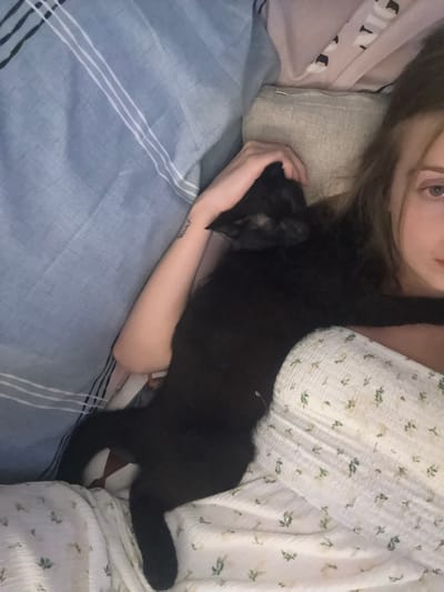 A young woman with long brown hair and light skin is lying down in bed, partially covered by blankets. A black cat is nestled comfortably against her. The woman is wearing a white top with a floral pattern. The bed appears to be in a bedroom. There is no indication of the specific location within Ratsada, Thailand, or any other identifying features of the environment.