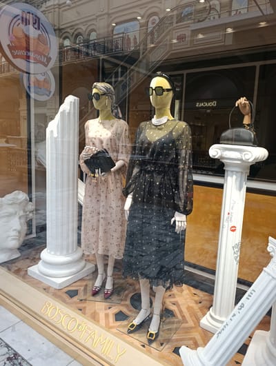 The image shows a window display featuring two mannequins dressed in fashionable attire. The mannequin on the left wears a light-colored, floral-print dress and holds a clutch purse. The mannequin on the right is dressed in a sheer black dress with a floral pattern and wears a pearl necklace. Both mannequins have sunglasses on. In the background, a classical sculpture of a head is visible, along with decorative columns. The flooring is a patterned wood. A sign with "BOSCO FAMILY" in illuminated yellow lettering is visible at the bottom of the display. The reflection in the window shows architectural elements of a building, including arches and a staircase, and signage. The overall impression is a high-end retail display. The location of this display is not explicitly indicated in the image, but if it were in Molokovo, Russia, it would likely be a boutique or department store.