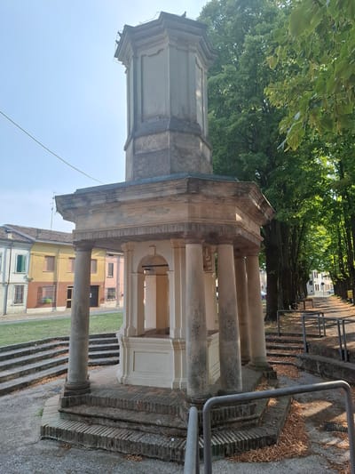 The image captures a historical structure, likely a monument or a public fountain, in Pieve Saliceto, Italy. The monument features a prominent octagonal base with columns supporting an upper structure. The material appears to be weathered stone and brick. In the background, there are residential buildings with colorful facades, including yellow and pink hues, and green shutters. A tree-lined pathway with benches runs along the right side of the image, suggesting a public park or street. The ground is paved, with fallen leaves scattered around, indicating an outdoor setting during autumn or late summer. A fence runs along the pathway. The sky is clear and bright. No people are visible in the image.