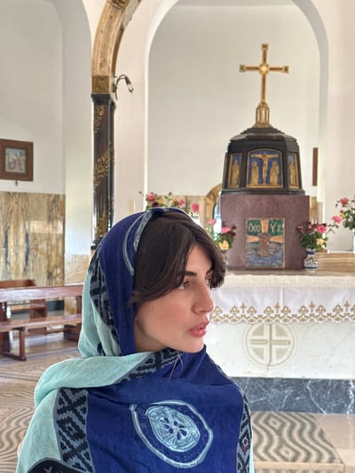 A person with dark hair, wrapped in a blue and light green patterned scarf, is looking upwards with their mouth slightly open. They are positioned in the foreground, with their head and upper body filling the right side of the frame. The background suggests an indoor setting, likely a place of worship. Visible elements include an altar with decorative panels and floral arrangements, a cross mounted on the wall, and ornate fabric with a patterned border. The overall ambiance appears serene and solemn, suggesting a moment of prayer or contemplation. The lighting seems to be artificial, indicative of an interior space. The location context provided places this scene within the Emek HaYarden Regional Council in Israel.