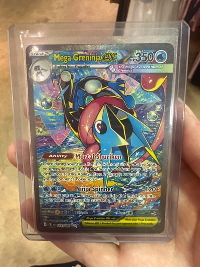 The image shows a person holding a Mega Greninja-EX Pokémon card in a protective sleeve. The card has a colorful, dynamic illustration of the Pokémon and text detailing its abilities and stats. The background is blurred, but appears to be indoors, possibly a room with shelves or objects. There are no discernible city landmarks or references to Halfway, United States of America.