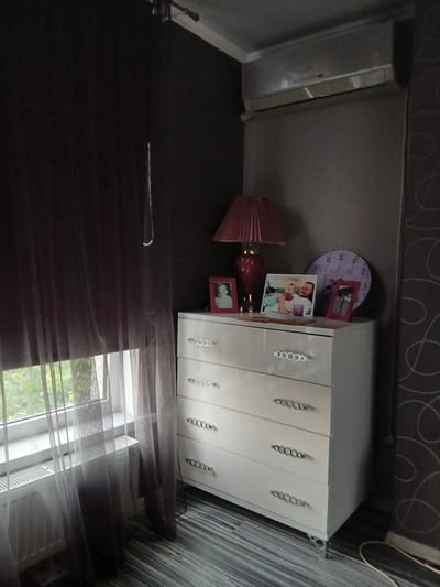 A white dresser with four drawers stands in a dimly lit room. Each drawer has two black, oval-shaped pulls. On top of the dresser are three framed photographs, a pink lamp with a pleated shade, and a round, purple clock with Roman numerals. A sheer, dark brown curtain hangs to the left of the dresser, partially obscuring a window. A white radiator is visible below the window. The room's walls are a dark color, one with a swirling pattern. An air conditioning unit is mounted high on the wall above the dresser. The floor is covered in dark, wood-patterned laminate. The scene appears to be an interior residential space, likely a bedroom, in Vita-Poshtova, Ukraine.
