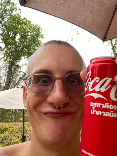 A close-up, low-angle shot captures a person holding a red Coca-Cola can next to their face. The person has light skin, short, wet hair, and wears glasses. A nose ring is visible. In the background, partially obscured by an umbrella, are trees and foliage. The location is Tambon Nam Phrae, Thailand, based on the text on the Coke can.