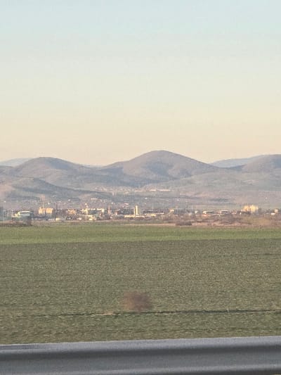 The image depicts a panoramic landscape overlooking the city of Diadovo, Bulgaria, under natural light conditions. In the immediate foreground, a blurred gray horizontal metal barrier, consistent with a guardrail, spans the bottom of the frame. Above this barrier, an expansive, flat, green agricultural field occupies the midground, extending towards the distant city. Faint horizontal striations are visible within the field, suggesting cultivation. A diffused, brownish, indistinct form is present in the lower-central portion of the field, possibly a reflection or distant vegetation.
Beyond the field, the intermediate background reveals the cityscape of Diadovo, characterized by a cluster of industrial and commercial buildings. Several structures exhibit prominent vertical elements, such as chimneys or towers, indicative of industrial activity. The city appears somewhat hazy due to distance.
The background consists of a series of rolling, dark gray-blue mountains or hills that rise above the city and extend across the horizon. The sky, occupying the upper portion of the frame, displays a subtle gradient, transitioning from a pale blue at the zenith to a light yellowish-orange hue near the mountain peaks, consistent with ambient daylight. No individuals, explicit actions, or interactions are discernible within the frame.
