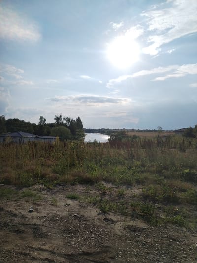 This media file depicts a serene, rural landscape under a bright, partly cloudy sky. The sun, high in the sky and creating a strong lens flare, suggests it is midday. The scene is a natural setting with a body of water, likely a lake or river, reflecting the light.

The foreground is dominated by dry, grassy terrain with some patches of sand and small stones. Tire tracks are visible on the ground, indicating recent vehicle activity. In the mid-ground, there are several houses with tiled roofs nestled among lush green trees. Further back, more trees and fields of dry grass stretch towards the horizon. The water body is a prominent feature, separating the vegetation on the left from the fields and buildings on the right.

The weather appears to be warm and fair, with scattered clouds adding texture to the bright blue sky. The overall atmosphere is peaceful and tranquil, evoking a sense of calm and quiet rural life. There is no visible text in the image. The location context provided is Moara Domnească, Romania, which aligns with the rustic, natural scenery.