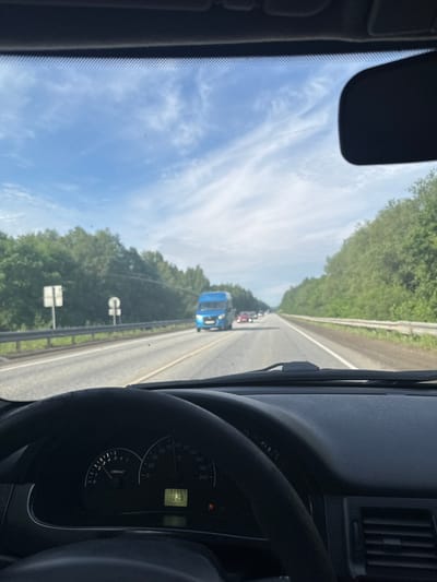 The image, taken from inside a vehicle, captures a driver's perspective of a road trip near Усть-Сыны, Russia. The scene looks forward through the windshield onto a highway bordered by dense green trees under a bright blue sky with scattered white clouds. The weather appears clear and sunny.

Ahead on the road, several vehicles are visible, including a prominent blue van, indicating light to moderate traffic. A road barrier runs along the right side of the highway. Partially visible road signs are too distant to read.

Inside the car, the right side of the frame shows a portion of the dashboard and the instrument cluster. The speedometer needle points to approximately 80-90 km/h, and the tachometer shows the engine running at around 2000-2500 RPM. A digital display on the cluster shows "190907", likely the odometer reading in kilometers, and "18:29", indicating the time is 6:29 PM. An orange warning light, possibly an airbag or seatbelt indicator, is illuminated on the cluster. A part of the rearview mirror is visible in the upper left corner of the frame. The overall impression is of a late afternoon or early evening drive on a highway through a forested area.