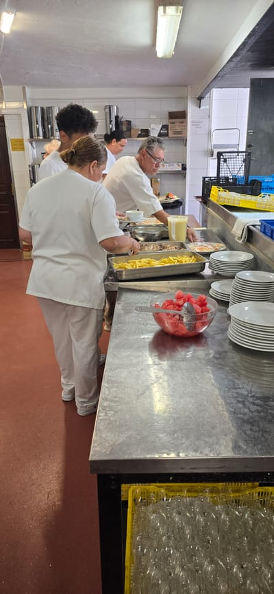 In a commercial kitchen, a group of individuals, likely kitchen staff, are engaged in food preparation and service. The scene is set in a brightly lit room with stainless steel countertops and white tiled walls.

In the foreground, a woman wearing a white chef's jacket and light-colored pants is seen from the waist down, actively working. In front of her, a metal tray holds a large portion of french fries. To the right of the french fries, a clear glass bowl is filled with diced watermelon, with a serving spoon resting inside. Further to the right, stacks of white plates are neatly arranged on the counter.

Behind the woman in the foreground, another individual, also in white chef attire, is visible from the neck up, focusing on their task. To the right of this person, a man in a white chef's coat and glasses is serving food from a tray. He appears to be placing an item onto a plate.

In the background, more kitchen equipment and supplies are visible. Several stainless steel beverage dispensers are lined up on a shelf, and various boxes and containers are also present. To the far right, a yellow plastic rack holds numerous wine glasses. The floor of the kitchen is a reddish-brown color. The overall impression is one of a busy, operational kitchen environment. The presence of the flag of Spain on a box in the background strongly suggests that this establishment is located in Spain. Given the context of the prompt specifying Sallent de Gállego, Spain, it is highly probable this image was taken in a kitchen within that municipality.