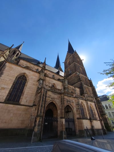 The photograph depicts the exterior of the Church of Saint Lawrence in Vysoké Mýto, Czechia, viewed from a low angle against a clear blue sky with scattered clouds. The church features Gothic architectural elements, including stone construction, pointed arches, spires, and ornate stonework. The facade is made of light-colored stone blocks. The sun is shining brightly, with its rays partially obscured by one of the church's spires. A cross with a figure of Christ is visible above one of the arched entrances, behind a black wrought-iron gate. In the foreground, a portion of a light-colored car and its antenna are visible. To the right of the church, another building with a white facade and numerous windows can be seen. Lush green foliage from a tree is also present in the upper right corner of the image. The ground in front of the church is paved with cobblestones.