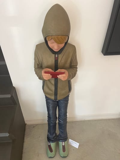 A full-body sculpture of a young boy is presented standing indoors against a plain white wall in Galatina, Italy. The sculpture depicts a boy with blond hair, wearing an olive green hooded jacket with dark grey and black trim, blue jeans, and light green shoes with dark accents. His head is bowed, and he holds a small, rectangular red object, resembling a smartphone, with both hands, positioned as if interacting with its screen. To the left, dark-colored, multi-tiered stairs are partially visible. To the right, a corner of a black-framed, dark triangular artwork hangs on the wall. The floor is light-colored. A white rectangular label, bearing text including 'GALATINA D'APRILE', 'SGUARDI E LUOGHI', and 'MOSTRA FOTO', rests on the floor near the sculpture's feet.