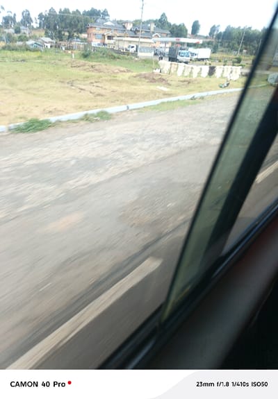 The image depicts an exterior view of Lari/Kirenya ward, Kenya, captured from the interior of a moving vehicle. The foreground shows a blurred, dark grey road surface alongside a light grey concrete curb with sparse green vegetation. A dark vertical vehicle pillar and window edge are visible on the right side. Beyond the curb, a large, dry, yellowish-brown grassy field occupies the midground. Within this field, multiple rows of white, rectangular objects, possibly sacks or fabrics, are laid out on the ground. Towards the right side of the midground, a fuel station is visible, marked by a "K-GAS" sign under a white canopy, with several vehicles, including a white truck and a white van, parked nearby. In the background, numerous multi-story buildings, some with dark roofs and light-colored facades (one prominent orange-brown), are nestled among dense green trees that extend to the horizon. The sky appears overcast. No discernible individuals or specific interactions are identifiable.