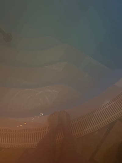 The image displays a close-up, downward angle of a person's feet standing at the edge of a jacuzzi or pool. The pool's water appears tinted with shades of blue and brown. The feet are positioned on a white, curved grating that lines the pool's edge. Water reflections and ripples are visible.