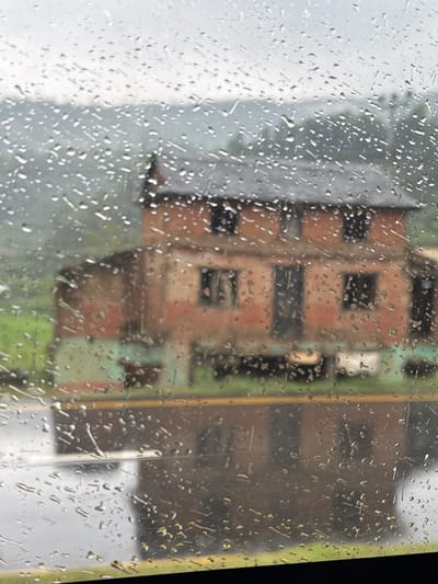 The image depicts a view through a rain-streaked window. Outside, buildings are visible, though obscured by the rain and the window's surface. One building appears to be made of brick with dark windows. The environment seems urban, though the specific location within Tharpu, Nepal, is not discernible. The primary subject is the rain on the window and the resulting distortion of the cityscape behind it.