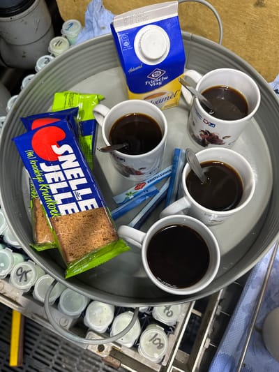 The image depicts a tray containing four mugs of coffee, two packets of "Snel Leven Kruidkoek" (spiced gingerbread cake), a carton of "Friesche Vlag" caramel half-and-half, and several packets of sugar. The tray is placed on a metal rack, and in the background, there are various small containers with lids, some labeled with numbers. A blue and white paper towel is also visible. The scene appears to be set indoors, possibly in a break room or staff area, given the presence of the rack with numbered containers which could be used for samples or ingredients in a laboratory or food production facility. The overall impression is one of a casual break or refreshment setup. The location in Wilhelminaoord, Netherlands is not evident from the visual information provided in the image.