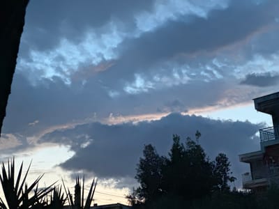 The image, taken from a low angle in Kalyves Polygyrou, Greece, shows a stormy sky at dusk. The clouds are a mix of gray and blue, with a small patch of lighter sky visible. In the foreground, on the left side of the image, there are silhouettes of yucca plants with spiky leaves. A dense bush or small tree is positioned in front of the clouds on the right side. A multi-story building, partially obscured by the tree, occupies the right side of the frame. The balcony railings of the building are visible. An electrical pole is visible behind the yucca plants.