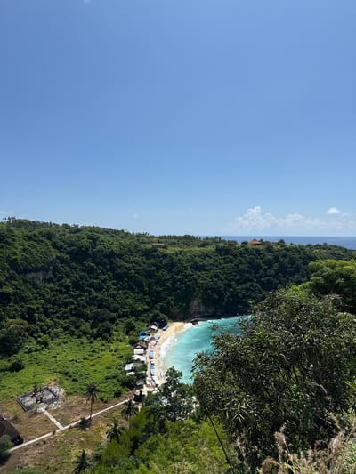 The image captures a coastal scene in Pejukutan, Indonesia. Lush, dense green vegetation covers the majority of the landscape, leading down to a beach with turquoise water. A small sandy beach is bordered by white-capped waves. Several structures, possibly huts or shacks, are situated near the shoreline, with a collection of colorful objects, likely beach umbrellas, arranged in a row along the beach. A path or walkway extends from the beach area toward an elevated point, where a structure or monument with dark-colored stone or concrete is situated. The sky is a clear blue with light clouds on the horizon. Trees and foliage are visible in the foreground, partially obscuring the view, suggesting the photograph was taken from a high vantage point, such as a cliff or elevated structure.