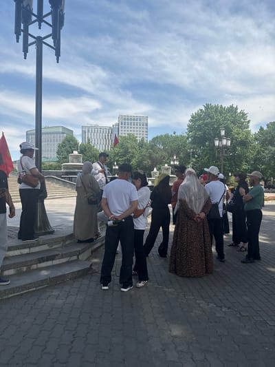A group of people stand along a sidewalk in Kashgar mahalla, Uzbekistan. The people are diverse in age and attire, with some wearing casual clothing, others in traditional dress. Some individuals hold a flag. They are positioned near a lamppost on a paved surface with trees, buildings, and a cloudy sky in the background. The setting is a public area, possibly a park or plaza.