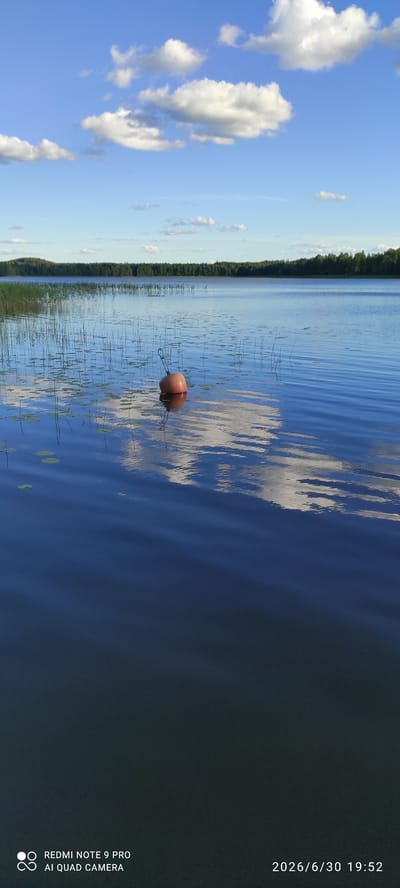A photograph captures a serene lake scene in Joutsa, Finland, likely taken on a sunny day. The image is oriented vertically, with the sky on the left and the water occupying the majority of the right side. White, fluffy clouds dot the bright blue sky, reflecting on the rippling surface of the lake.
A single, orange buoy floats on the calm water, tethered by thin ropes that disappear into the lake's depths. Surrounding the buoy are sparse reeds and lily pads, indicating a shallow area near the shore. The distant shoreline is lined with a dense forest of green trees, suggesting a natural, unpopulated setting. The overall mood of the image is peaceful and tranquil.
Visible text in the bottom right corner reads "2026/6/30 19:52" and "REDMI NOTE 9 PRO AI QUAD CAMERA". This indicates the date and time the photo was taken, as well as the device used. The lighting suggests it is late afternoon or early evening, given the time stamp.