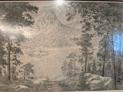 This is a black and white etching depicting a landscape scene. The foreground features a grassy, rocky area with several trees and bushes. In the middle ground, there is a body of water, possibly a lake or river, with a few small islands. The background shows a densely forested hillside leading up to a large, prominent rock formation that resembles a layered pyramid. Beyond that, distant mountains are visible, with atmospheric perspective suggesting a hazy or misty environment. The style of the etching is detailed, with fine lines used to render the textures of the trees, rocks, water, and foliage. The overall impression is one of a serene, natural wilderness. There is no visible text in the image. The location context of Ankara, Turkey, does not provide any specific cues within the artwork itself to confirm or deny this location. The time of day and weather are not explicitly indicated but the lighting suggests daytime.