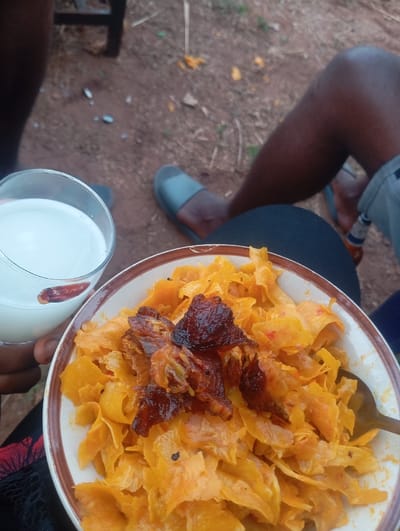 A bowl of what appears to be cooked plantains, topped with dark, roasted meat pieces, sits on a person's lap. A glass of white liquid, possibly milk, is held alongside. The scene is outdoors, evidenced by the ground and legs of people wearing casual footwear. A beer bottle is partially visible in the background.