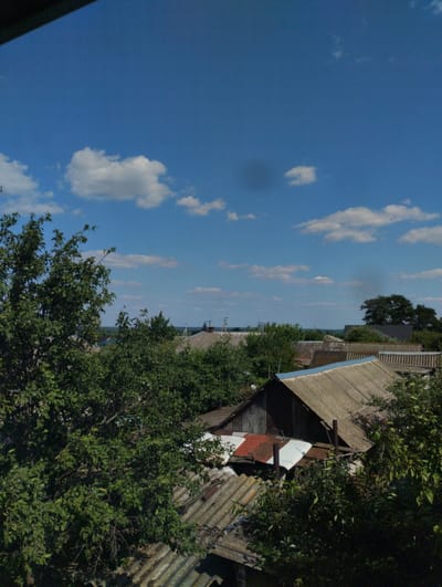 The image captures a panoramic view of a residential area, likely in the city of Shchuchye, Russia, under a bright blue sky with scattered white clouds. The foreground is dominated by lush green trees and foliage, partially obscuring the view of several houses. These houses have corrugated, weathered roofs, some showing signs of age and repair, with one having a distinct red and white metal roof. In the background, more houses and trees are visible, extending towards a distant horizon that suggests a flat landscape. The overall impression is of a rural or semi-rural setting during daytime.