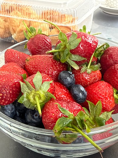 A close-up shot depicts a clear plastic container filled with strawberries and blueberries, positioned near a partially visible plastic container containing baked goods. The berries appear fresh and ripe. The scene appears to be indoors, possibly in a kitchen or dining area. There is no information to confirm this image was taken in the city of Olginskaya, Russia.
