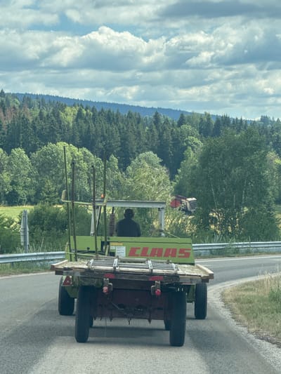 A person is driving a green CLAAS agricultural vehicle with a trailer full of hay down a paved road in Longchaumois, France. The vehicle is on the right side of the road. The road is surrounded by trees and fields. The sky is cloudy with some patches of blue. The image appears to have been taken from another vehicle on the road.