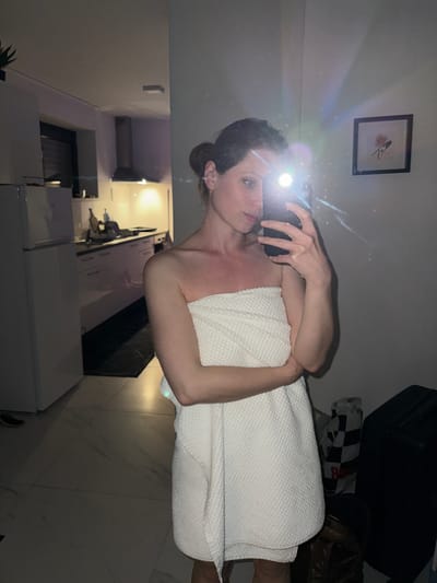 A woman wrapped in a white towel takes a selfie with a phone flash obscuring her face. The scene is indoors, likely a bathroom or bedroom adjacent to a kitchen. Visible in the background are kitchen countertops, cabinets, and appliances. There is no information to suggest the location is in Kranj, Slovenia. The picture above shows a picture on the wall, a black suitcase, and a black-and-white checkered shopping bag.