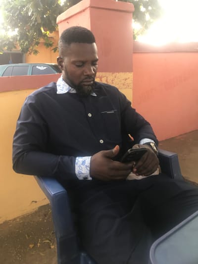 A male subject with dark skin, a short beard, and a high-fade haircut is seated outdoors in Ugwolawo, Nigeria. He occupies a blue plastic chair, looking downwards at a dark-colored smartphone held with both hands, appearing engaged with its screen. He wears a navy blue long-sleeved shirt featuring a white collar and patterned white and light blue cuffs, along with white buttons and a pen slot on the left chest pocket. A gold-toned watch is present on his left wrist. The background consists of a two-toned wall, with an orange-red section to the subject's right and a yellow-brown section to his left. A reddish pillar is situated behind his head. Green leafy vegetation is visible above the yellow-brown wall, and a portion of a green vehicle is discernible in the far left background. The ground is composed of light brown soil, and a dark, reflective surface, likely a table, is partially visible in the bottom right foreground.
