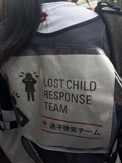 The image displays a close-up, rear view of an individual in the city of Kanazawa, Japan, wearing a specialized vest. The person has long, dark hair visible over their left shoulder and down their back. They are wearing a white upper garment with a small red embroidered symbol, possibly a double horizontal bar, visible at the collar area. Over this, a dark, mesh-textured vest is worn, featuring a large transparent plastic pocket on the back. A black strap from a backpack or bag is visible on the right shoulder.
Inside the clear pocket of the vest, a white sheet of paper is inserted, displaying text and an illustration. The prominent English text reads "LOST CHILD RESPONSE TEAM" in black capital letters. To the left of this text is a black silhouette of a child figure, depicted with hands covering its face and tear-like droplets around its head, suggesting distress. Below the English text, a red horizontal line is present, followed by a red cross symbol. Adjacent to the cross, Japanese text reads "迷子捜索チーム" (Maigo Sōsaku Chīmu), which translates to "Lost Child Search Team." A second red horizontal line appears below the Japanese text. A smaller, faint white text, unidentifiable, is visible near the red cross. The overall appearance suggests a uniform for personnel involved in locating missing children.