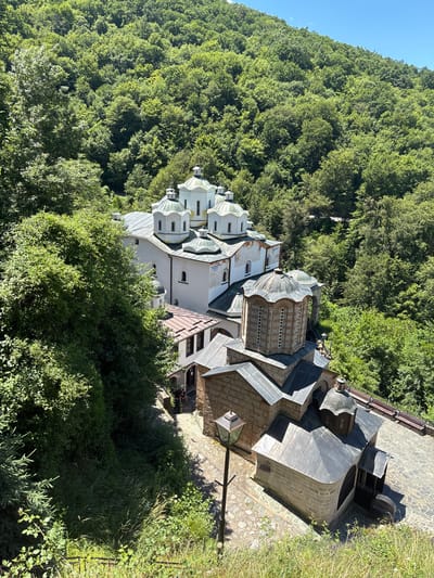 This aerial, high-angle, close-up shot depicts the Monastery of Saint Jovan Bigorski, a historic Orthodox Christian monastery located in the vicinity of the city of Kriva Palanka, North Macedonia. The monastery complex is nestled within a dense, green forest on a hillside. The main church, with its distinct white facade and multiple green domes, is a prominent feature. Adjacent to it are smaller, stone buildings with darker, sloped roofs. A cobbled pathway winds through the complex, and a lamppost stands near one of the stone structures. Lush vegetation, including trees and bushes, surrounds the monastery. The sky is clear and blue. Two individuals are visible sitting on a bench near an entrance to one of the buildings, and another person is standing near the same area.