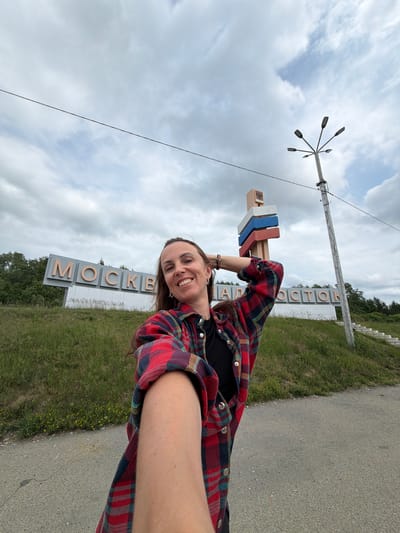 A woman stands on a paved road, taking a selfie. She is wearing a red and blue plaid shirt over a black t-shirt and has her left arm raised, with her hand behind her head. She is smiling and looking at the camera.

Behind her, a large sign with the word "MOSCOW" in large, metallic letters is visible. To the right of the sign, a lamppost with multiple lamps extends upwards, and a smaller sign with Russian flag colors is attached to a pole. The environment is grassy with some yellow wildflowers. The sky is cloudy, with patches of blue visible. The road surface is asphalt.