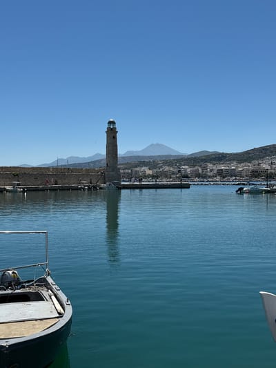 The image depicts the Venetian harbor of Rethymno, Crete, Greece, on a clear, sunny day. In the foreground, a small, gray motorboat is moored, its bow extending into the lower-left quadrant of the frame. The boat has a weathered appearance with visible wear and tear. The turquoise water of the harbor is calm, with gentle ripples reflecting the bright blue sky.
Dominating the mid-ground is the iconic Venetian lighthouse, a tall, stone structure with a distinctive lantern room at its apex, situated at the end of a long, stone quay. The quay is lined with people strolling and several small boats are docked along its edge. The stone fortifications of the harbor wall are visible, with a weathered, ancient appearance.
In the background, the city of Rethymno unfolds, a dense collection of buildings with white and earth-toned facades. Behind the city, a range of mountains rises, with one prominent peak appearing somewhat conical and distant. The overall scene conveys a sense of a peaceful, sun-drenched coastal town.