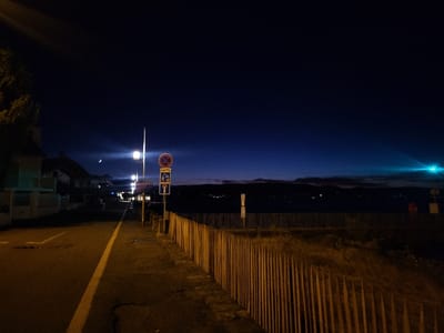Night scene. A paved road with a yellow lane marker runs along a wooden fence bordering a dark body of water. A row of streetlights illuminates the road and adjacent buildings. A crescent moon is visible in the dark blue sky. A road sign with a red circle and other symbols stands along the road. The water appears to stretch to a distant shoreline with twinkling lights.