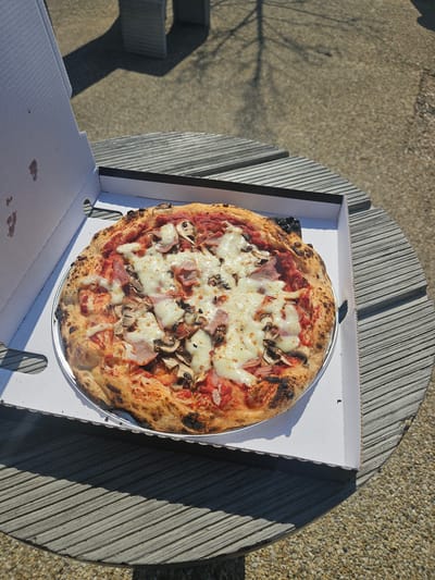 A freshly baked pizza, appearing to be a ham and mushroom variety (Reine), rests on a silver pan inside an open white cardboard delivery box. The pizza features a well-browned crust with some charring, a tomato sauce base, melted white cheese, sliced mushrooms, and pieces of ham. The interior of the box lid shows minor brown stains. The pizza box is situated on a round, ridged grey outdoor table. Beyond the table, a light-colored gravel or aggregate ground surface is visible. The scene is illuminated by strong sunlight, casting distinct shadows from the table and a tree (indicated by a branch shadow pattern on the ground). No individuals are present. The location is an outdoor setting within the city of Fréjus, France.