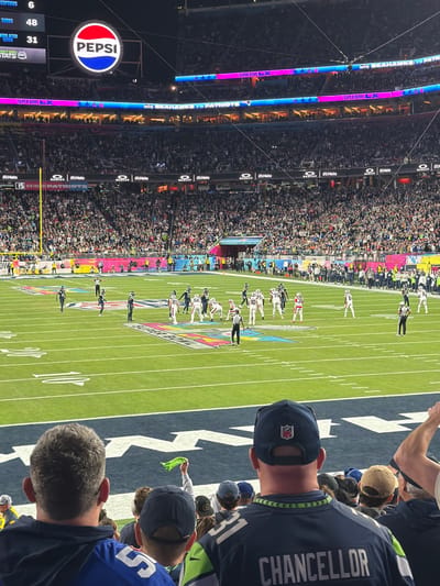 The image captures an American football game, likely the Super Bowl, from a fan's perspective. A Pepsi advertisement is visible on a screen. The score indicates the Los Angeles Rams leading, 48-31. The field is marked with "Super Bowl" logos, and a player wearing a "Chancellor" jersey is in the foreground. Spectators fill the stadium, and the game is in progress. A combination of players in white and blue uniforms are set up for the play on the field.