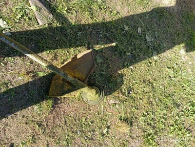 An outdoor scene in Урыцкі сельскі Савет, Belarus shows a string trimmer lying on a grassy area under a partial shadow. The trimmer, predominantly yellow, rests with its shaft angled across the frame from the upper left to the lower center. The cutting head, a dark circular housing, is visible at the lower center, containing coiled string and surrounded by freshly cut grass. The ground is covered with a mixture of green and dry grass, with some sparse weeds and fallen leaves scattered throughout. A shadow, cast by an unseen object, falls across the scene from the upper right, creating a distinct contrast with the sunlit areas. In the upper left corner, a piece of weathered wood is partially visible, lying on the grass. The overall impression is of a recently used or temporarily abandoned lawn maintenance tool in a rural setting.