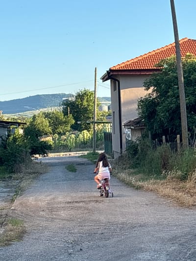 In Enevo, Bulgaria, a young girl with long dark hair, wearing a white t-shirt and pink shorts, rides a small pink bicycle with training wheels down a gravel road. The road is flanked by overgrown grass and bushes. To the right, a light-colored house with an orange tiled roof is visible, partially obscured by a large tree. A tall utility pole stands near the house. In the distance, rolling hills and more trees form the background under a clear blue sky. The scene appears to be a rural residential area during daylight.
