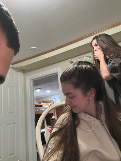 In Ocean City, United States of America, three individuals are present in a room. One person, partially visible on the left, has dark hair and is positioned in the foreground. In the center, a young woman with long brown hair is seated and appears to be having her hair clipped. She is wearing a light-colored hoodie. Behind her, another young woman with long brown hair is visible, covering her mouth with her hand. The background reveals a doorway leading to another room, which contains a bunk bed and wooden furniture. The lighting suggests an indoor setting, possibly a residential area.